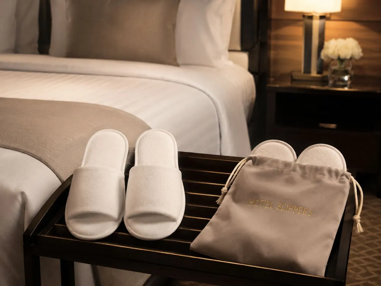 Custom-branded hotel slippers by UMYAN with a drawstring pouch beside a guest bed